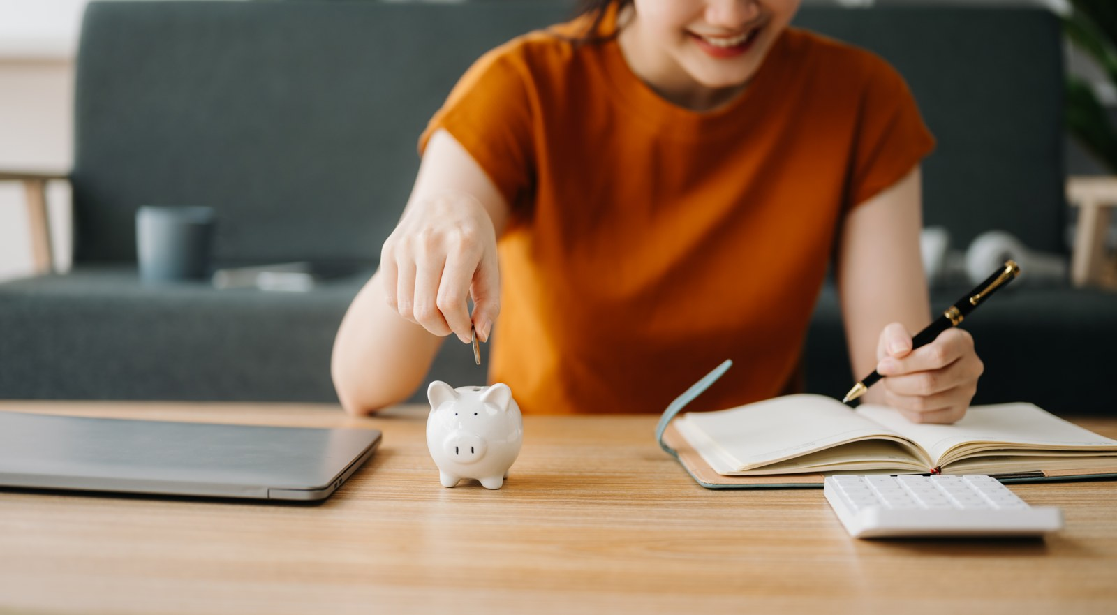 A woman putting a coin into a piggy bank beside a notebook