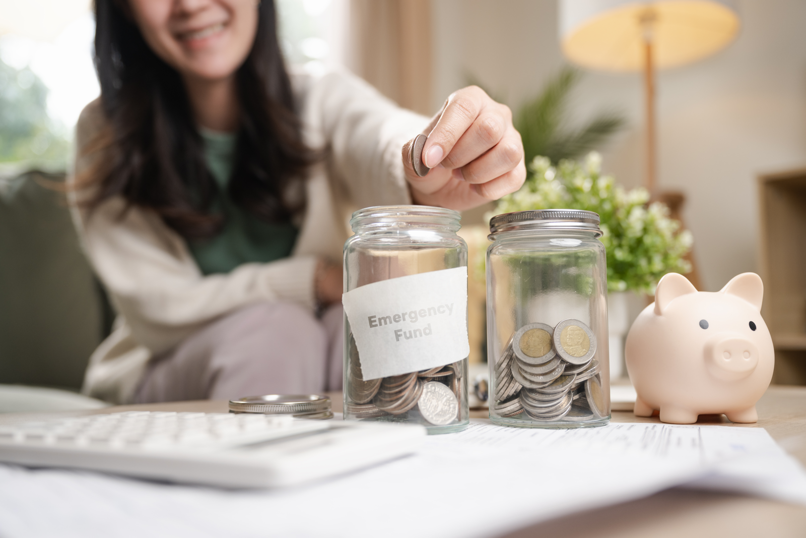 A woman adding a coin to an emergency fund jar beside savings tools